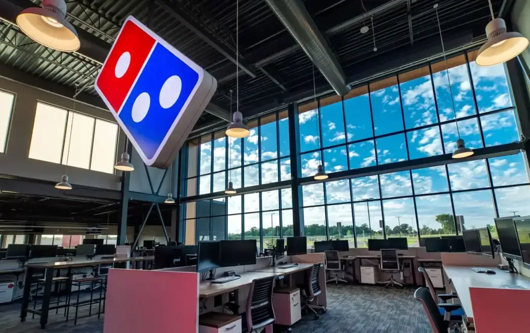 Open office space with large windows and workstations. A lighted sign with a red and blue domino is prominently displayed, hanging from the ceiling.