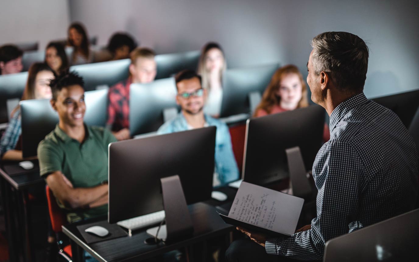 A teacher stands at the front of a classroom, facing students seated at desks with computers. The students appear attentive, with one holding a notebook.