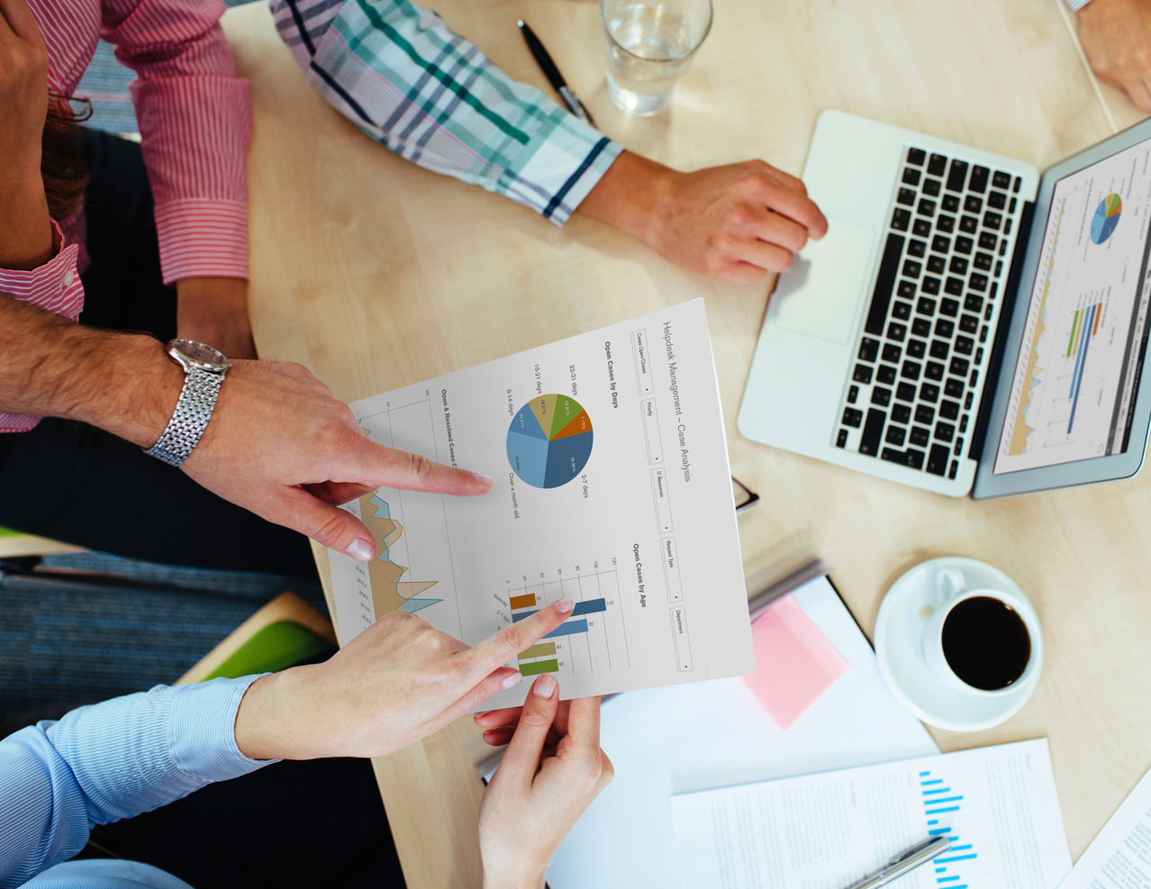 People are sitting at a table reviewing a document with graphs and charts. There is a laptop, coffee cup, and various office supplies on the table.