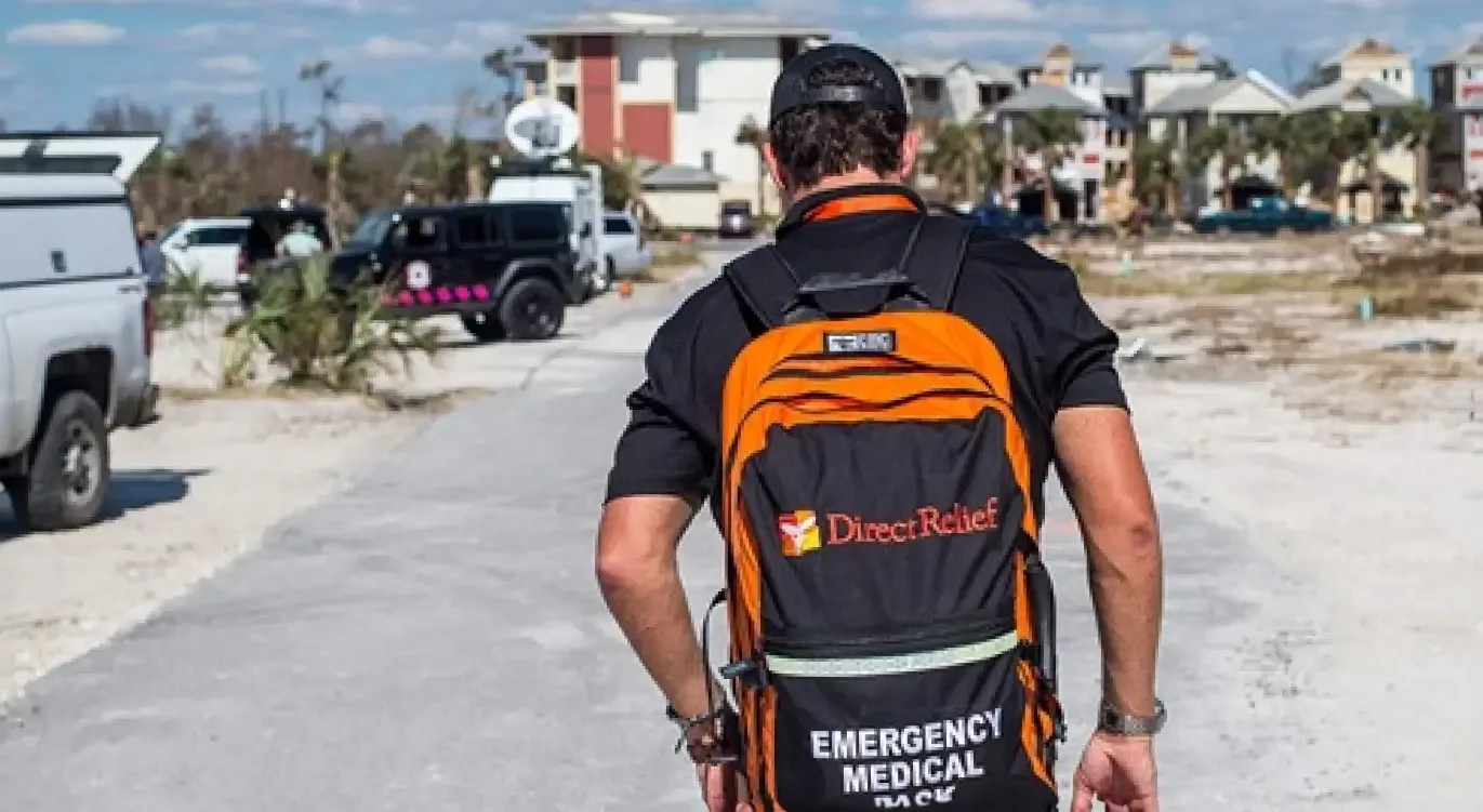 A person wearing an "Emergency Medical Pack" backpack with the Direct Relief logo walks towards a disaster-stricken area with damaged buildings and emergency vehicles in the background.