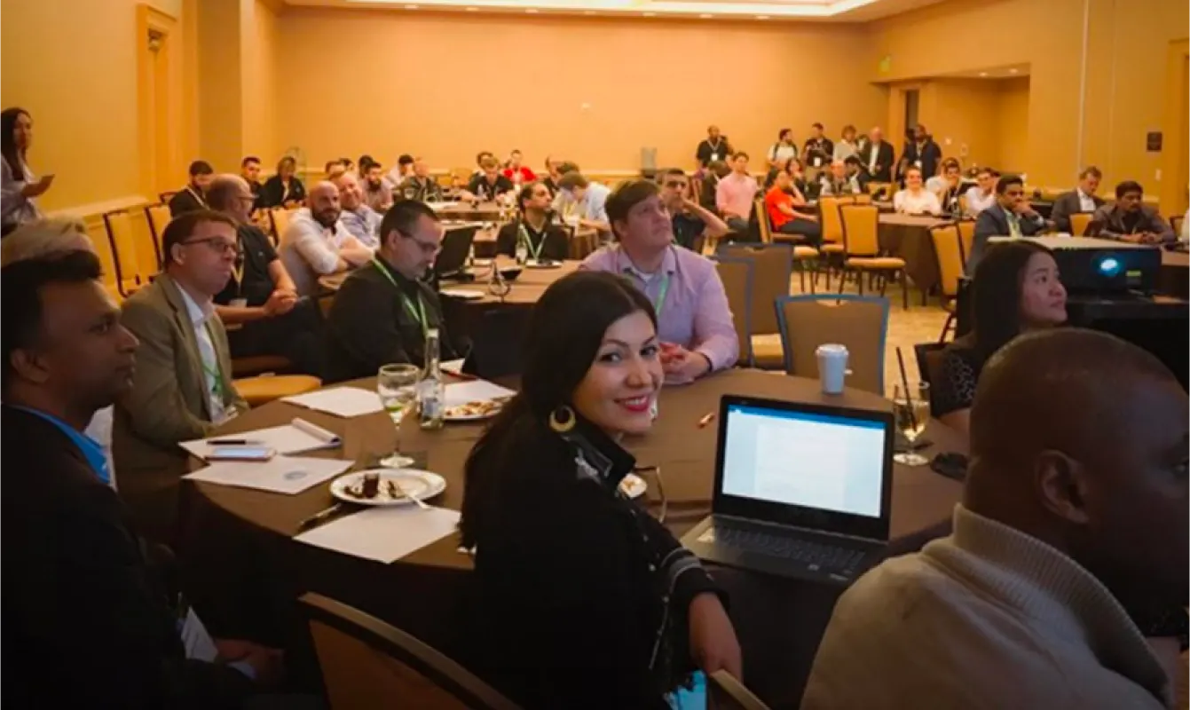 A diverse group of people sits attentively at round tables during a conference, some with laptops and documents in front of them, in a large, well-lit room.