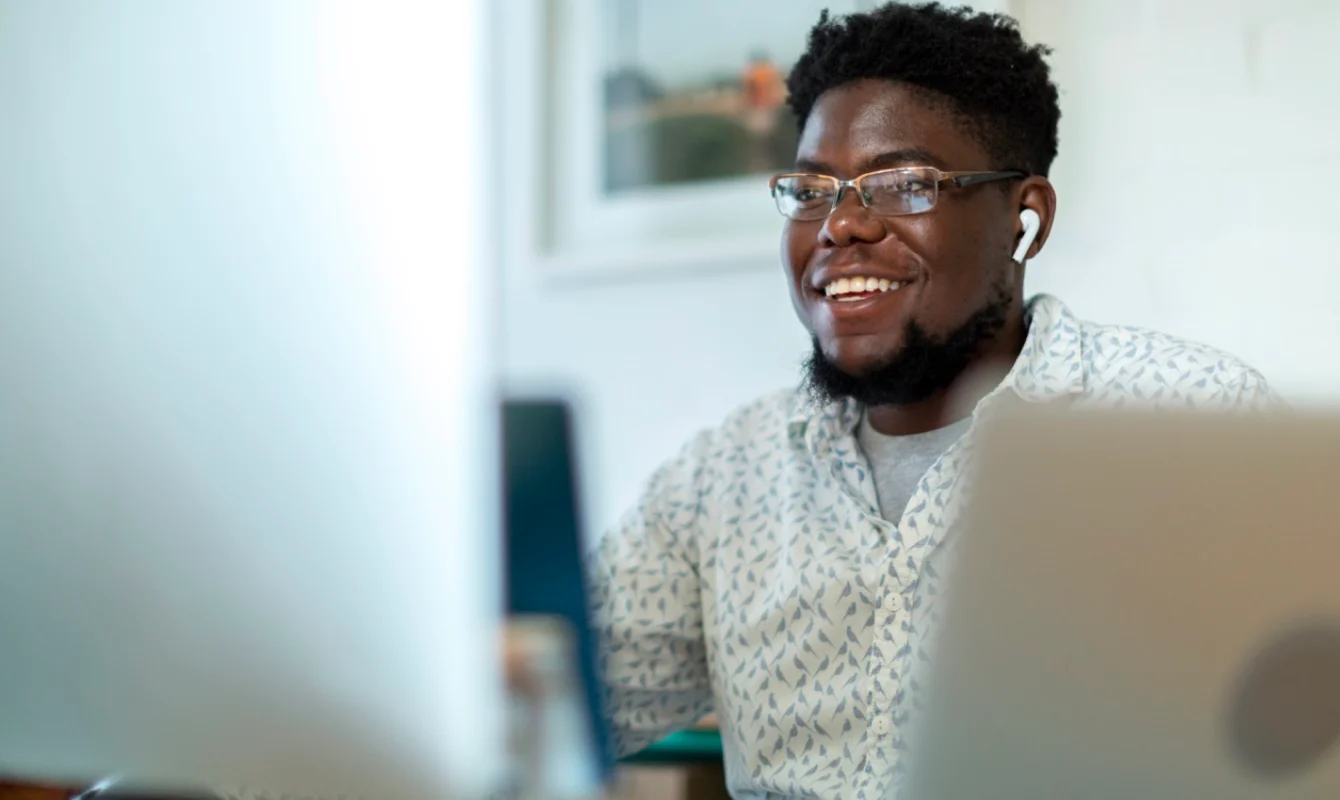 A person wearing glasses and earphones is smiling while using a laptop, with another screen visible in the foreground.