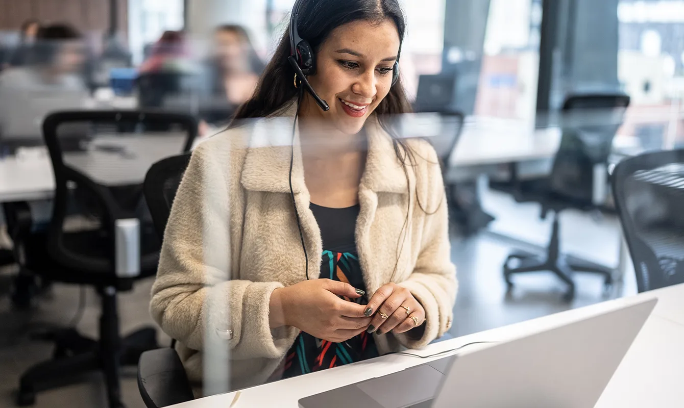 A woman wearing a headset and light-colored jacket sits at a desk with a laptop, working in an office with other blurred individuals in the background.