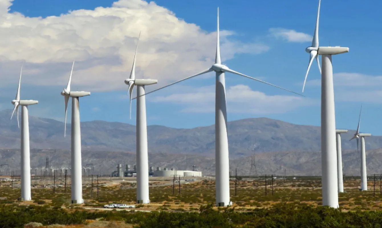 Wind turbines in a desert landscape, with mountains in the background and a few buildings visible in the distance.