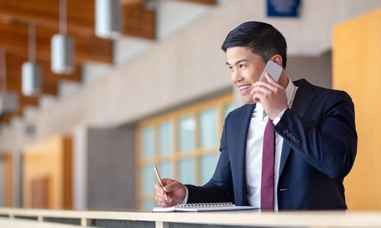 A man in a suit and tie talks on a phone and writes in a notebook while standing at a counter in an office setting.