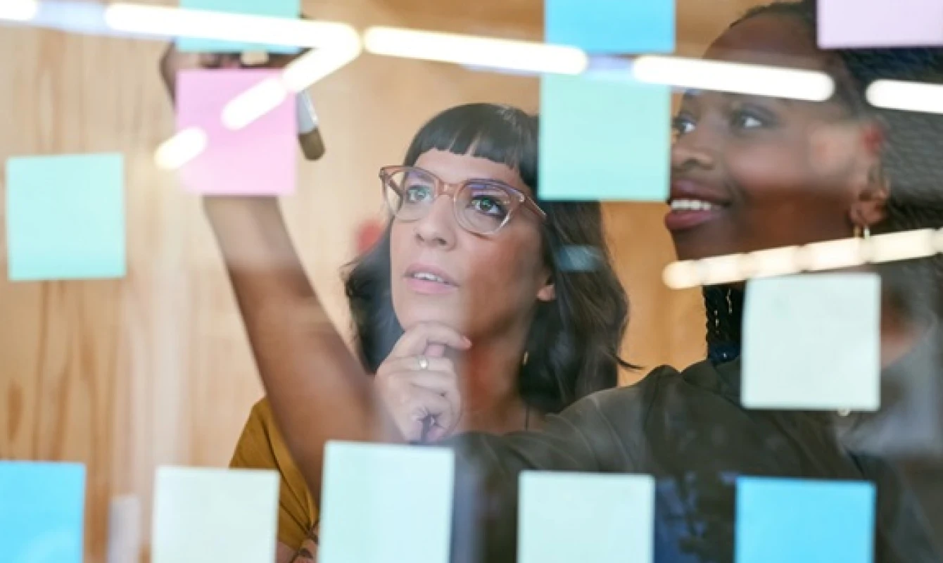 Two women are collaborating, writing on a glass board filled with colorful sticky notes in an office setting.