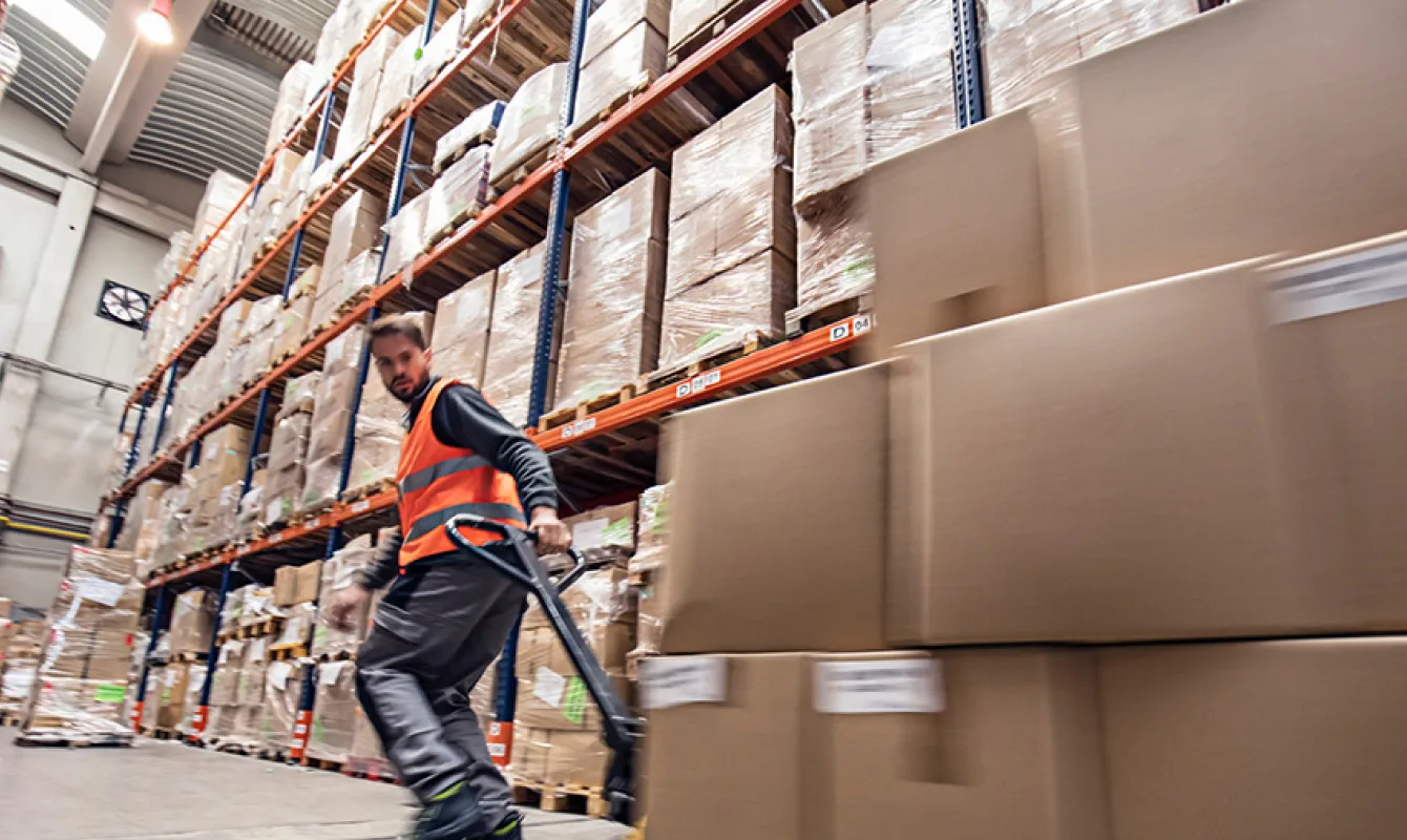 A warehouse worker in an orange vest moves large boxes with a hand pallet truck in an aisle surrounded by tall shelving units stacked with cardboard boxes.