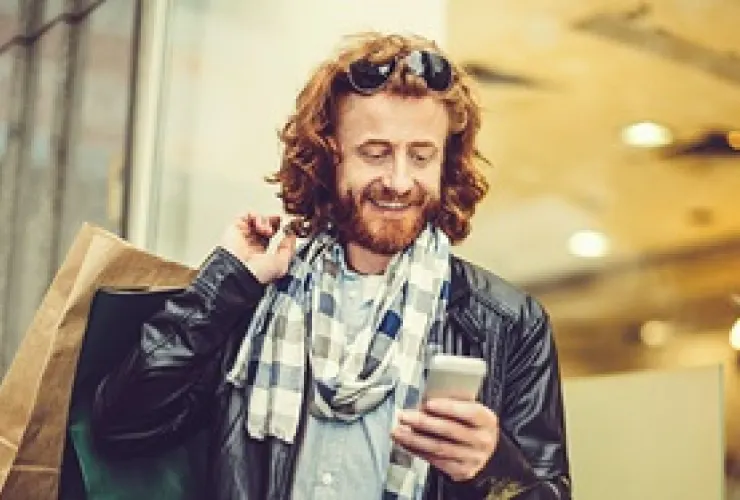 A bearded person with wavy red hair, wearing sunglasses, a scarf, and a leather jacket, holds shopping bags over their shoulder and looks at their phone while smiling.